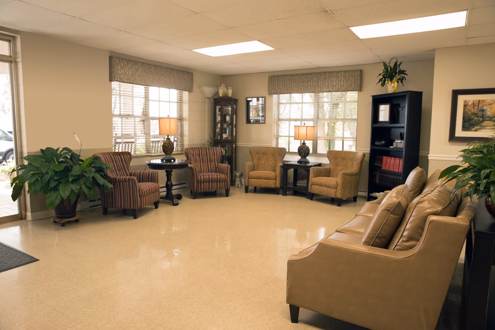 A well-lit living room area in a senior living facility featuring a tan leather sofa, two striped armchairs, two beige armchairs, side tables with lamps, a tall black bookshelf, a glass display cabinet, potted plants, and large windows with blinds and valances.