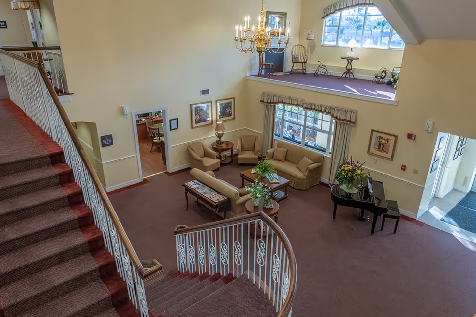 View from the top of a carpeted staircase looking down into a spacious living room area with beige sofas, armchairs, a coffee table, a side table with a lamp, a black grand piano with a flower arrangement, and large windows letting in natural light. A chandelier hangs from the ceiling, and there is a small elevated alcove with chairs and a table near a window.