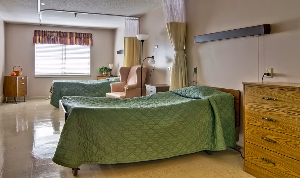A room with two single beds covered with green quilts, a pink armchair between the beds, a wooden dresser on the right, a small wooden cabinet on the left, and a window with blinds and a valance letting in natural light.