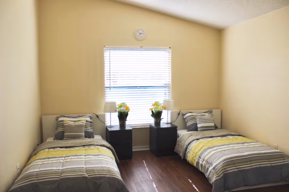 A simple bedroom with two single beds placed parallel to each other against opposite walls. Each bed has striped bedding in shades of gray, white, and yellow, with matching pillows. Between the beds is a window with closed blinds, flanked by two black nightstands, each holding a lamp and a vase with yellow flowers. A small clock is mounted on the wall above the window. The walls are painted beige and the floor is wooden.