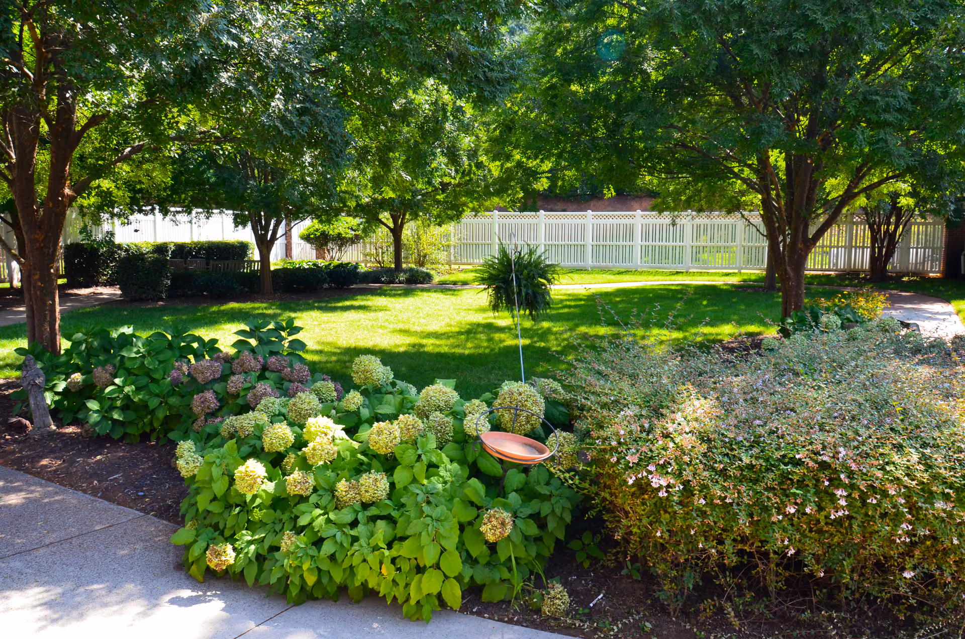 A sunny outdoor garden area with lush green grass, flowering bushes, and several trees providing shade. A white picket fence encloses the garden, and a concrete pathway runs along the edge of the flower beds.