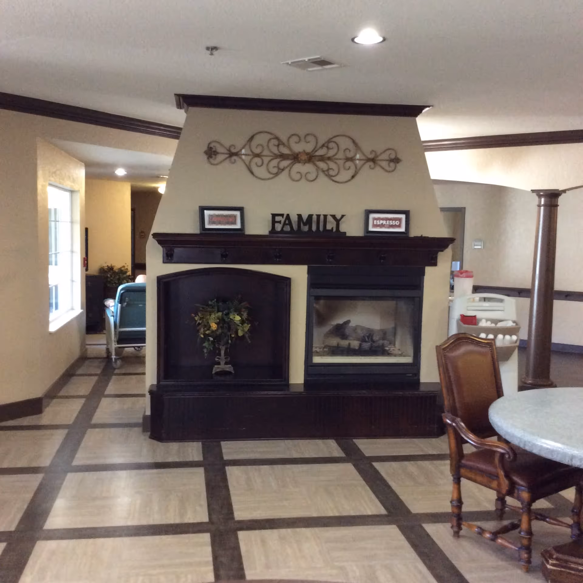 Interior common area with a fireplace and decorative shelf displaying the word FAMILY and two framed pictures labeled CAPPUCCINO and ESPRESSO. There is a round table with a wooden chair in the foreground, a window on the left, and a hallway with a wheelchair visible in the background.