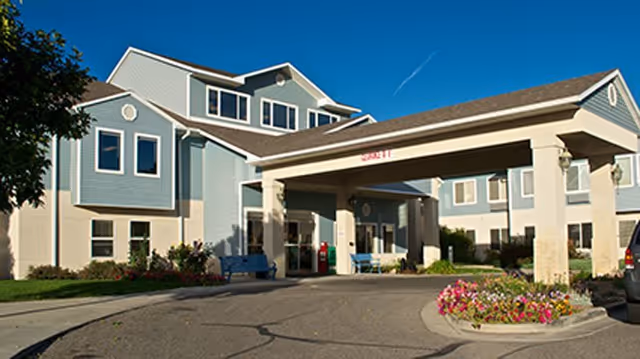 Exterior view of a senior living facility building with a covered entrance driveway, blue and beige siding, multiple windows, and landscaped flower beds near the entrance.