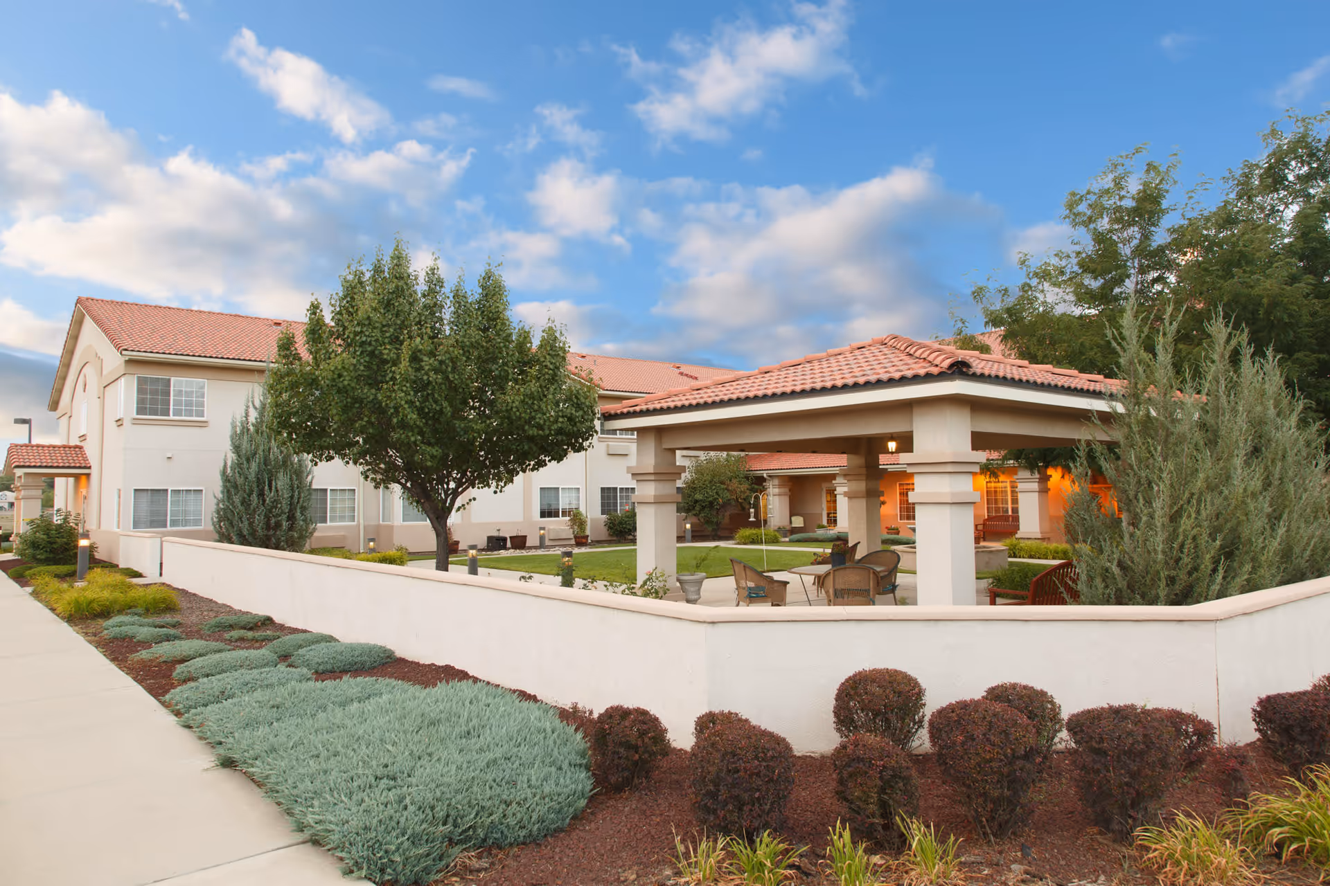 Outdoor courtyard area of The Bridge at Farmington Assisted Living facility featuring a covered seating area with chairs and tables, surrounded by landscaped bushes, trees, and a low white wall under a partly cloudy blue sky.