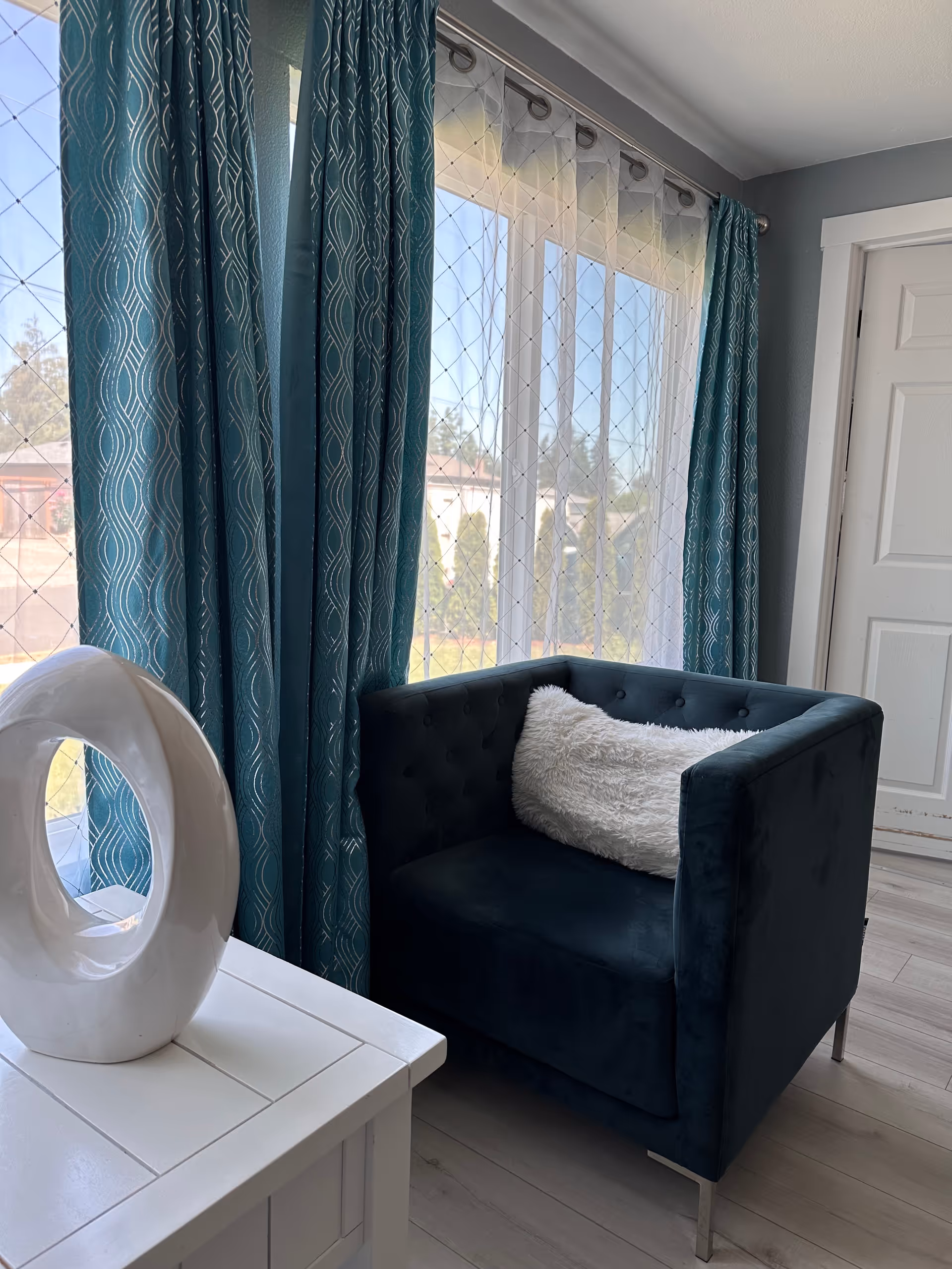 A cozy corner of a room featuring a dark teal upholstered armchair with a white fluffy pillow. Next to the chair is a white table with a modern white circular sculpture on top. The background shows large windows with sheer white curtains and teal patterned drapes, allowing natural light to brighten the space. A white door is visible on the right side.