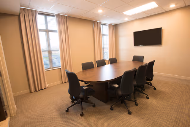A small conference room with an oval wooden table surrounded by eight rolling office chairs, two tall windows with curtains, and a wall-mounted TV.