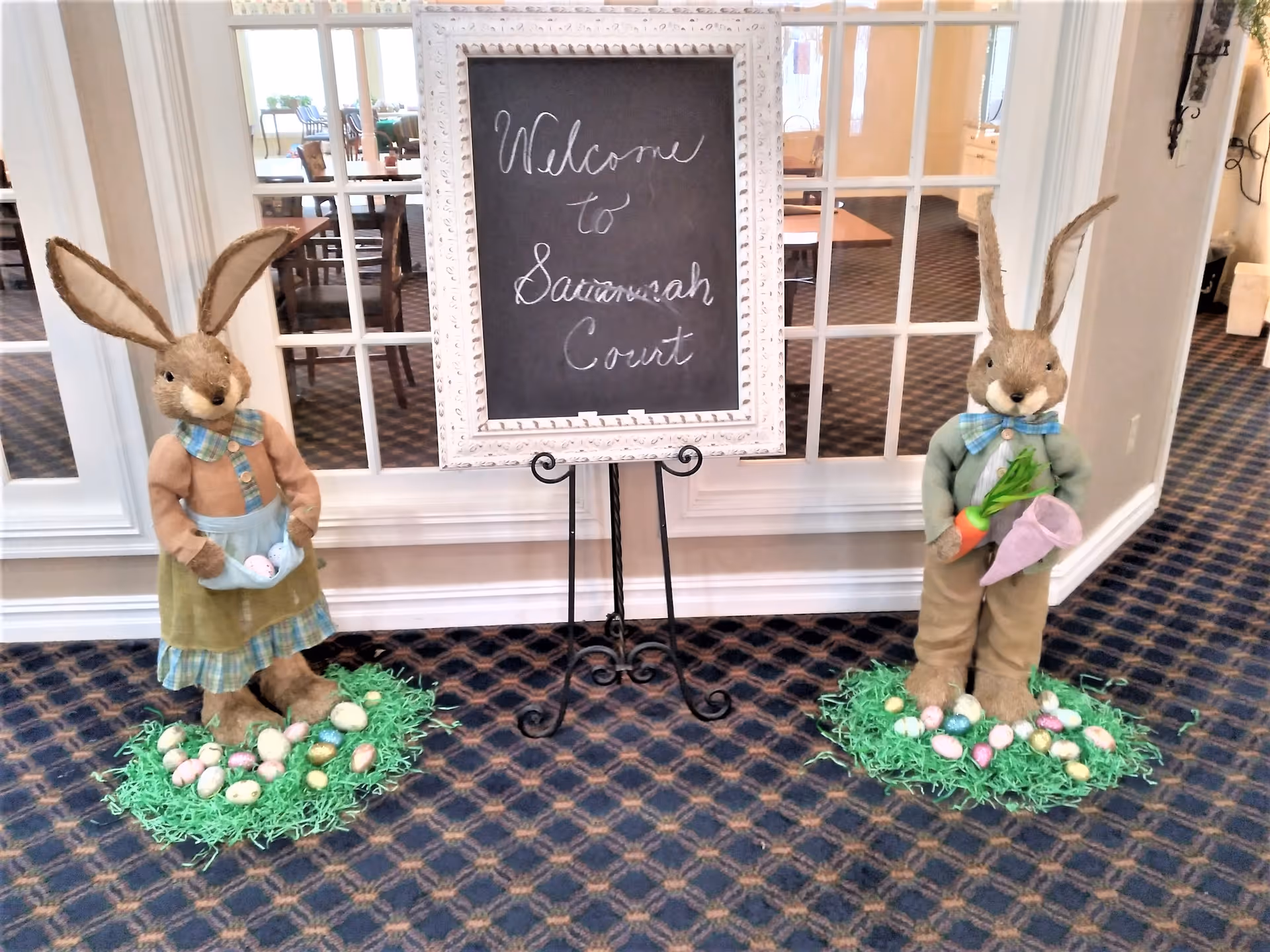 Two large decorative bunny figures standing on green artificial grass with colorful Easter eggs scattered around. Between them is a white-framed chalkboard on a black metal stand with the handwritten message 'Welcome to Savannah Court'. The background shows a room with tables and chairs visible through glass-paneled doors.