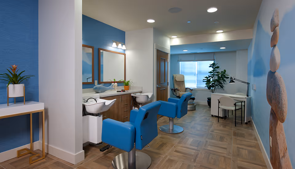 Interior view of a salon area in a senior living facility with two blue salon chairs facing mirrors and wash basins, a beige pedicure chair near a window with blinds, a small desk with a white chair and lamp, a plant, and a wall mural of stacked stones on the right.