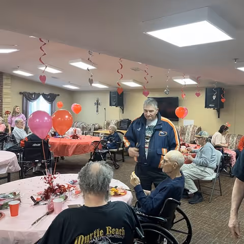 Seniors and visitors gathered around decorated tables with pink and red balloons and heart streamers in a community dining/activity room.