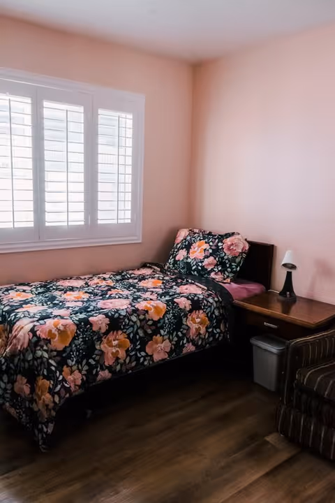 A small bedroom with a single bed covered in a floral patterned comforter and matching pillow. The walls are painted light pink, and there is a window with white shutters on the left side. Next to the bed is a wooden nightstand with a small lamp and a trash bin underneath. Part of a striped armchair is visible on the right side.