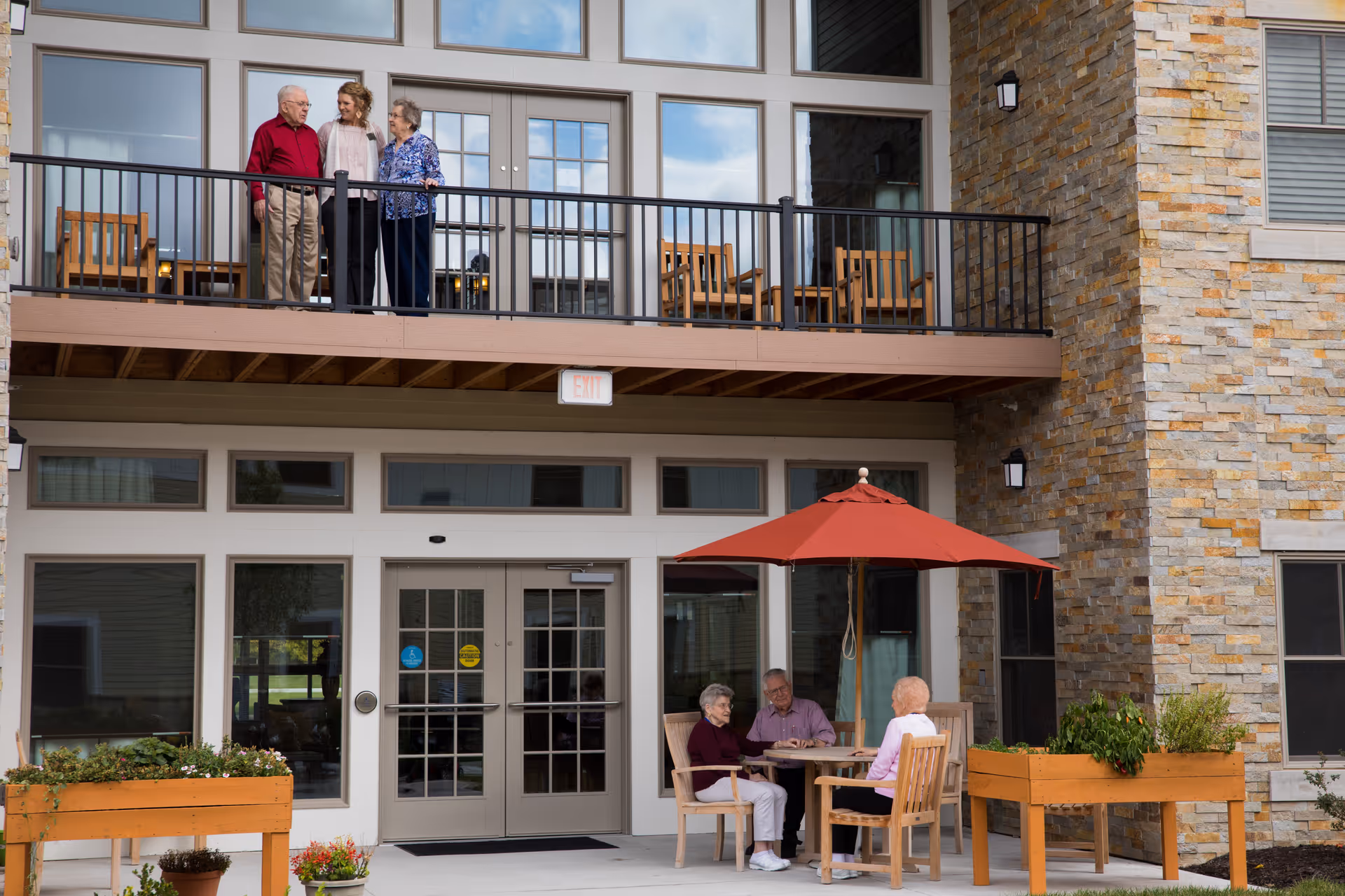Three elderly people and a younger woman standing and talking on a balcony outside a building, while three elderly people sit at a table with a red umbrella on a patio below, surrounded by planters and a stone wall exterior.