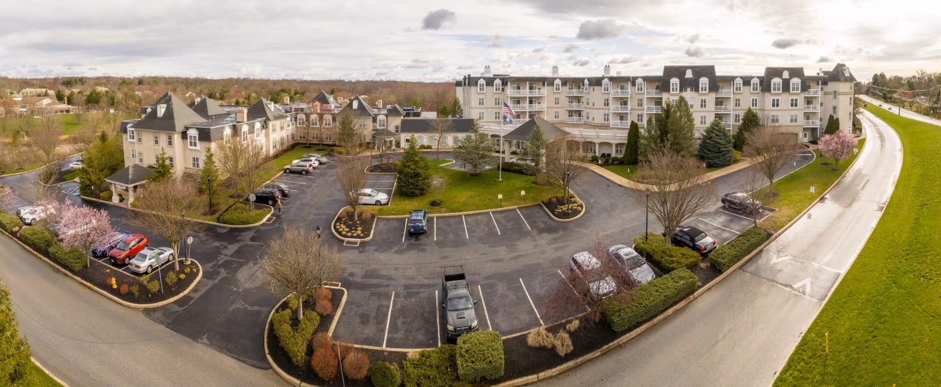 Aerial view of Wellington at Hershey's Mill senior living facility showing a large multi-story building with a parking lot in front, landscaped greenery, trees, and a road curving around the property under a cloudy sky.