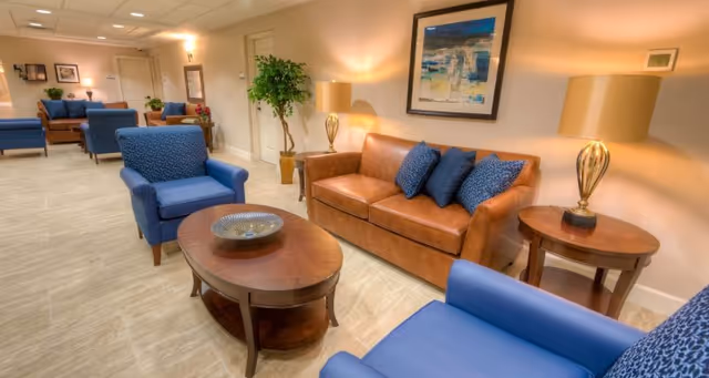 A cozy living room area with a brown leather sofa adorned with blue cushions, two blue armchairs, a wooden oval coffee table with a decorative bowl, and two wooden side tables each with a lamp. The walls are light-colored and decorated with framed artwork and a large green plant in the corner. The room has a warm and inviting ambiance with soft lighting.