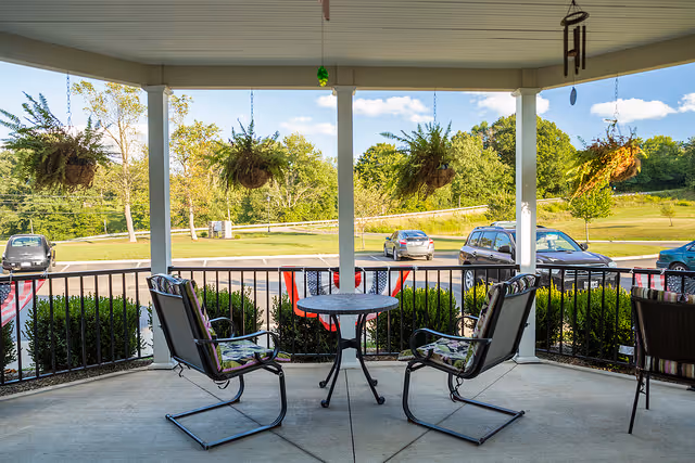 Covered outdoor patio area with a round table and four chairs, two of which have floral cushions. Hanging plants are suspended from the ceiling, and a wind chime is visible on the right side. Beyond the patio, there is a parking lot with several cars and a grassy area with trees under a blue sky with some clouds.
