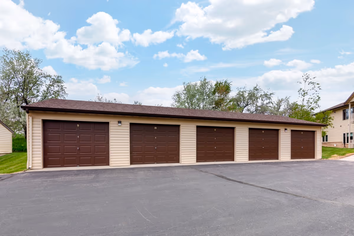 A row of five brown garage doors numbered 9 to 13 on a beige building with a brown roof, situated on a paved driveway with green grass and trees in the background under a partly cloudy blue sky.