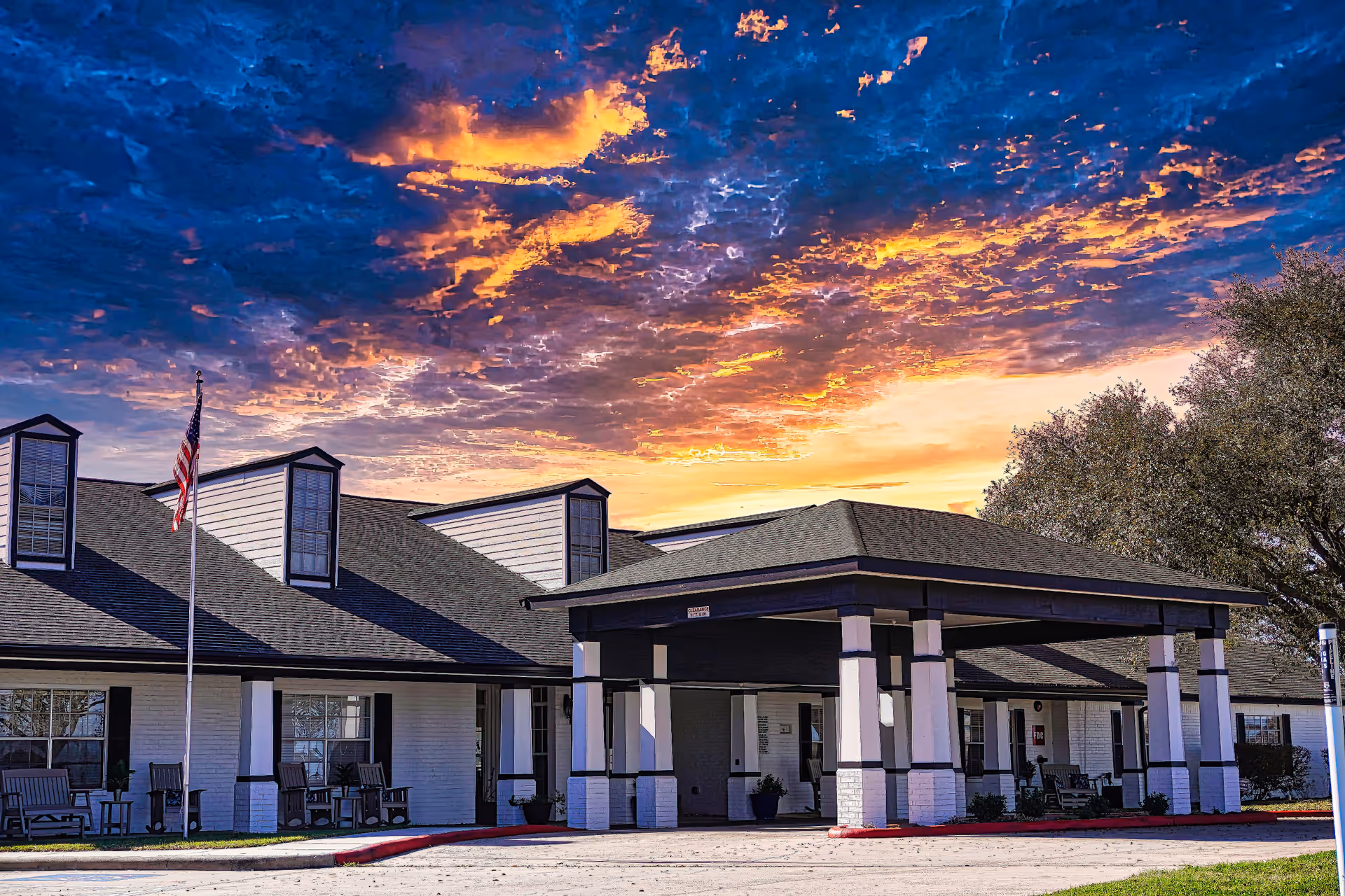 Exterior view of Sodalis Victoria Assisted Living building at sunset with a dramatic sky filled with orange and blue clouds. The building has a covered entrance supported by white pillars with black trim, rocking chairs on the porch, and an American flag on a flagpole.