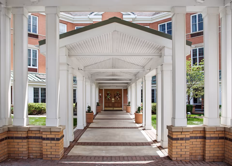 Covered white-columned walkway leading to double wooden entrance doors of a brick senior living building.