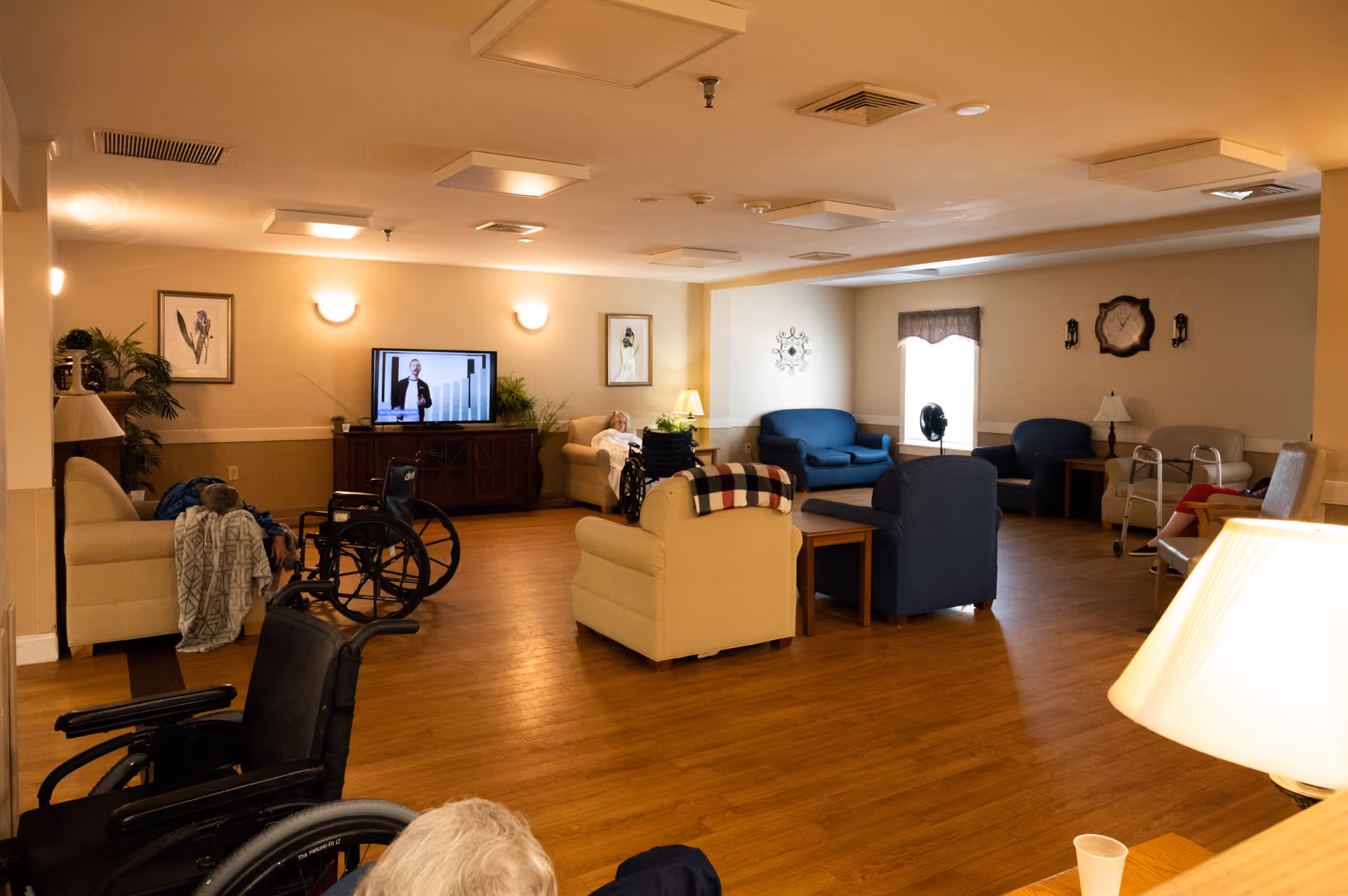 A spacious senior living common area with several armchairs and sofas arranged around a television mounted on a wooden cabinet. The room has wooden flooring, soft lighting from wall sconces and lamps, framed artwork on the walls, and a few wheelchairs and walkers are visible. Two elderly individuals are seated in the room, one in a wheelchair and another reclining on a sofa.