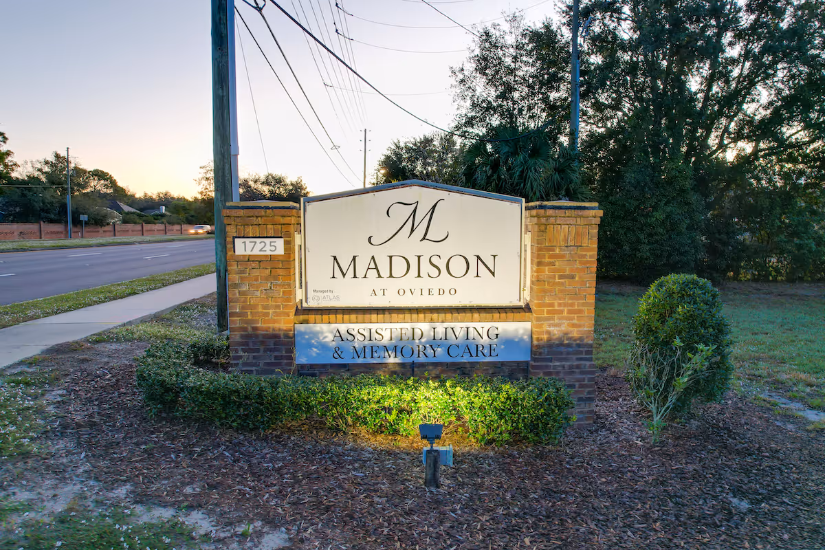 A brick and stone sign for Madison at Oviedo, an assisted living and memory care facility, situated next to a road with trees and bushes around it during early evening.