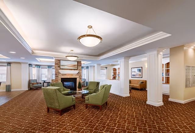 A spacious senior living facility common area with a patterned brown carpet, four green upholstered armchairs arranged around a glass coffee table, and a stone fireplace with a landscape painting above it. The room has white columns, recessed lighting, large windows with curtains, and additional seating areas with chairs and a sofa.
