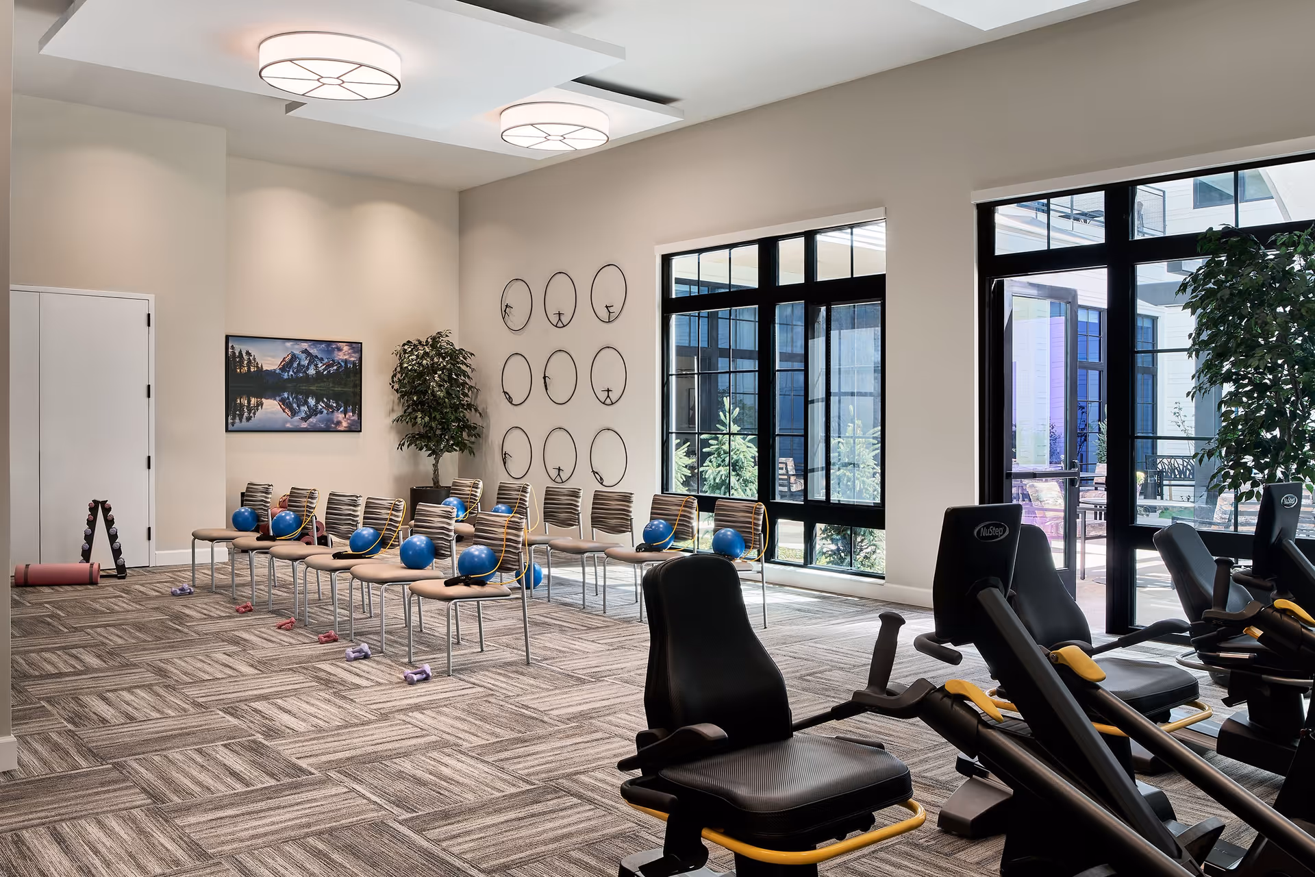 A bright fitness room with exercise chairs, blue exercise balls on chairs, small dumbbells on the floor, and large windows letting in natural light. The room has modern ceiling lights, a potted plant, and a wall-mounted TV displaying a mountain landscape.