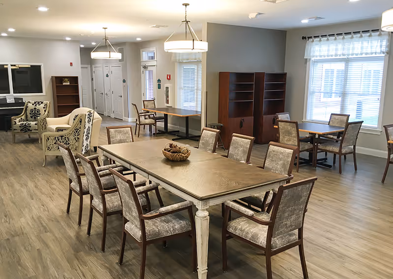 A spacious, well-lit common area with multiple tables and chairs arranged for group seating. The room features wood flooring, large windows with blinds, two pendant lights, and a few armchairs near a piano in the background. There are also wooden bookshelves and a door leading to another area.