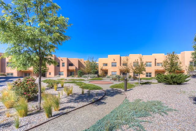 Outdoor courtyard area at The Montecito of Santa Fe featuring a winding gravel pathway, desert landscaping with small trees, bushes, and ornamental grasses, and a two-story adobe-style building in the background under a clear blue sky.
