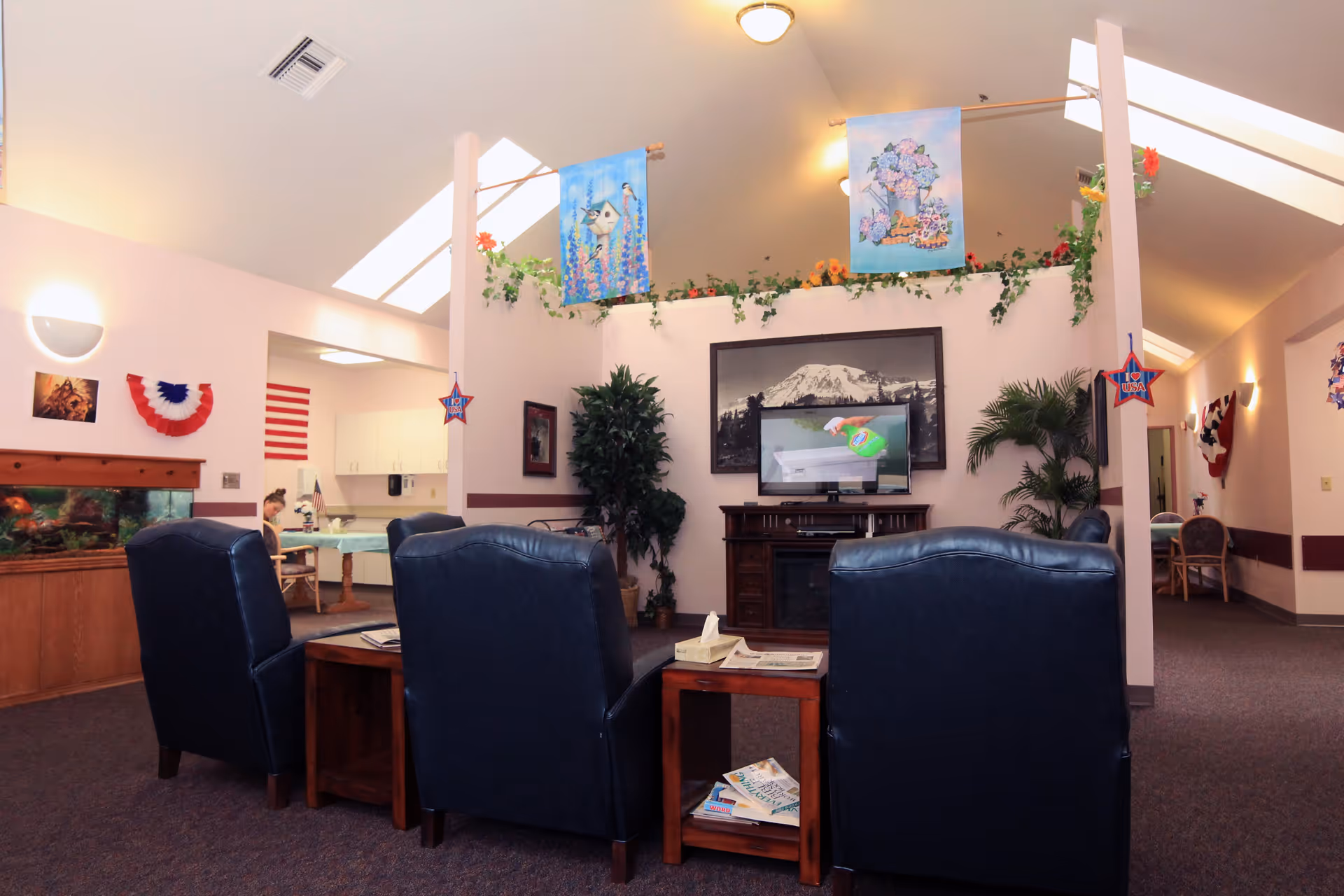 A cozy living room area in a senior living facility with three dark blue armchairs arranged around a wooden coffee table. The room features a large black and white photo of a mountain above a TV on a wooden stand, potted plants, and decorative banners hanging from the ceiling. In the background, there is an open kitchen area and a hallway with additional seating. The space is decorated with patriotic and floral-themed decorations.