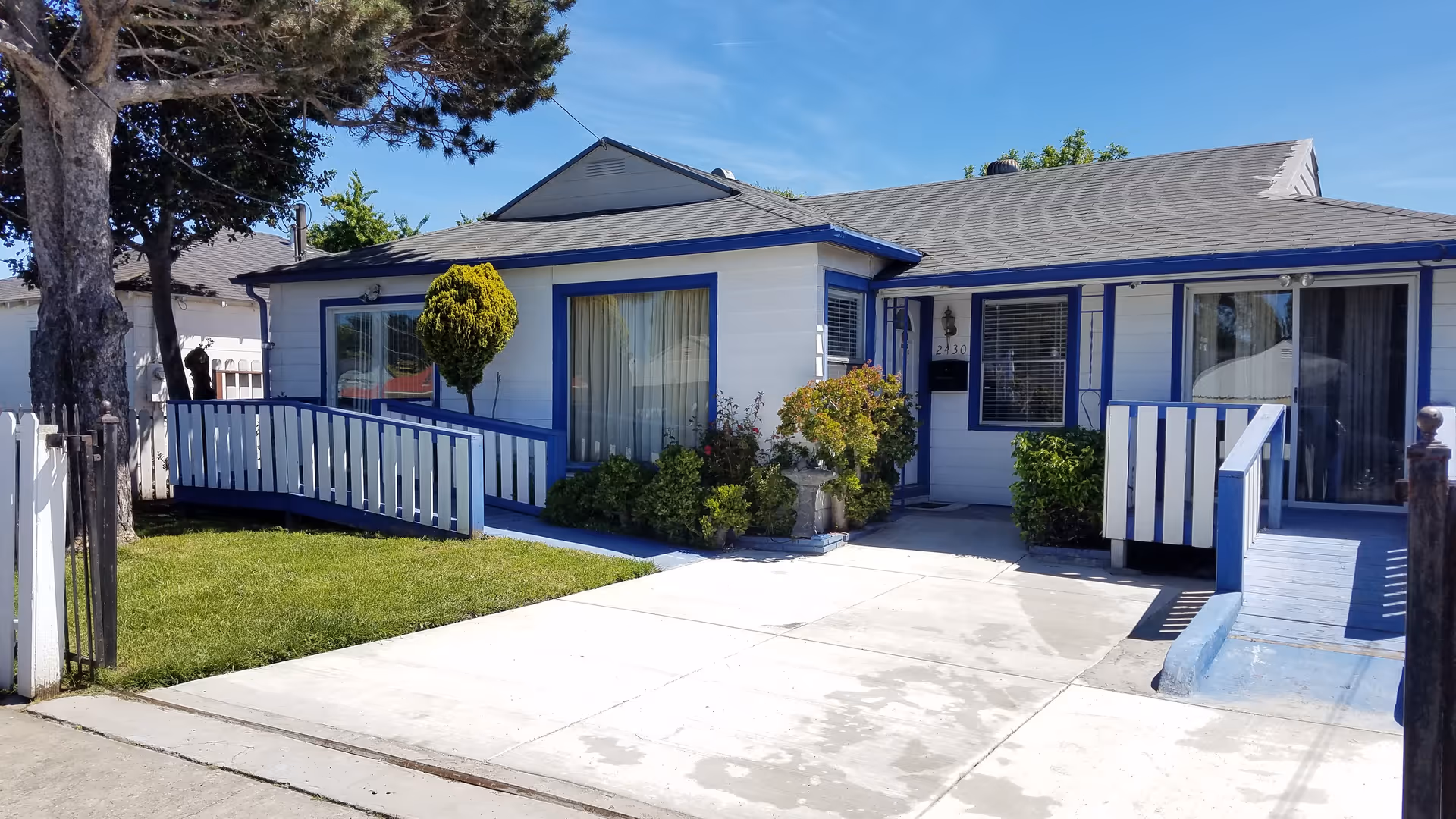 Exterior view of a single-story residential building with white walls and blue trim, featuring two wheelchair ramps leading to separate entrances. There is a small lawn with bushes and a tree on the left side, and a concrete driveway in front under a clear blue sky.