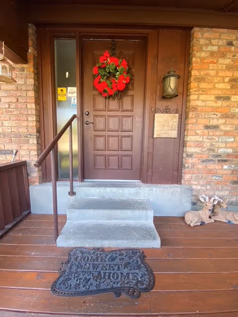 Entrance porch showing a brown front door with a red wreath, concrete steps and handrail, a 'Home Sweet Home' doormat, and brick siding.