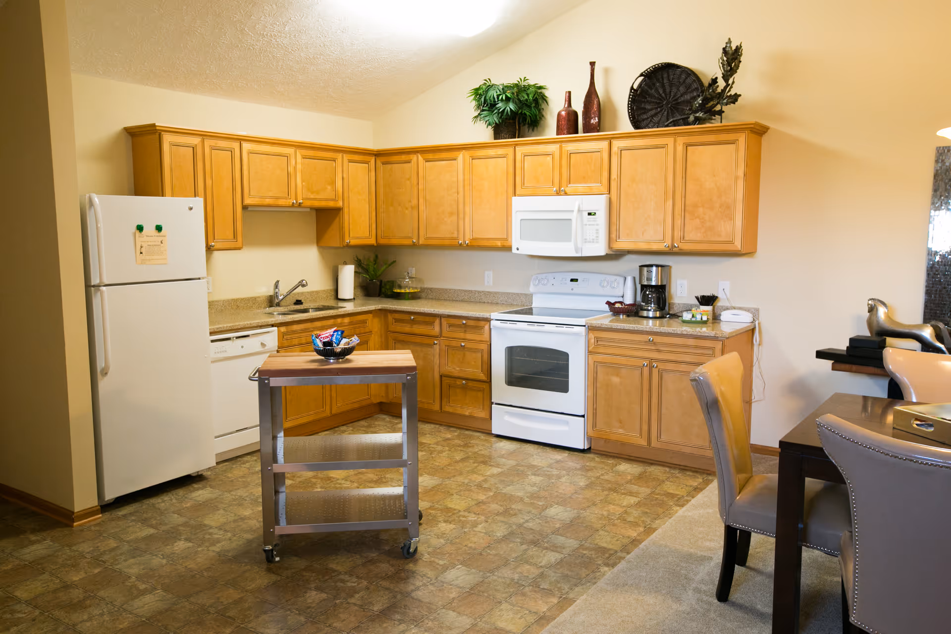 Bright kitchen with light wood cabinets, white refrigerator and stove, a small rolling island cart, and adjacent dining chairs.