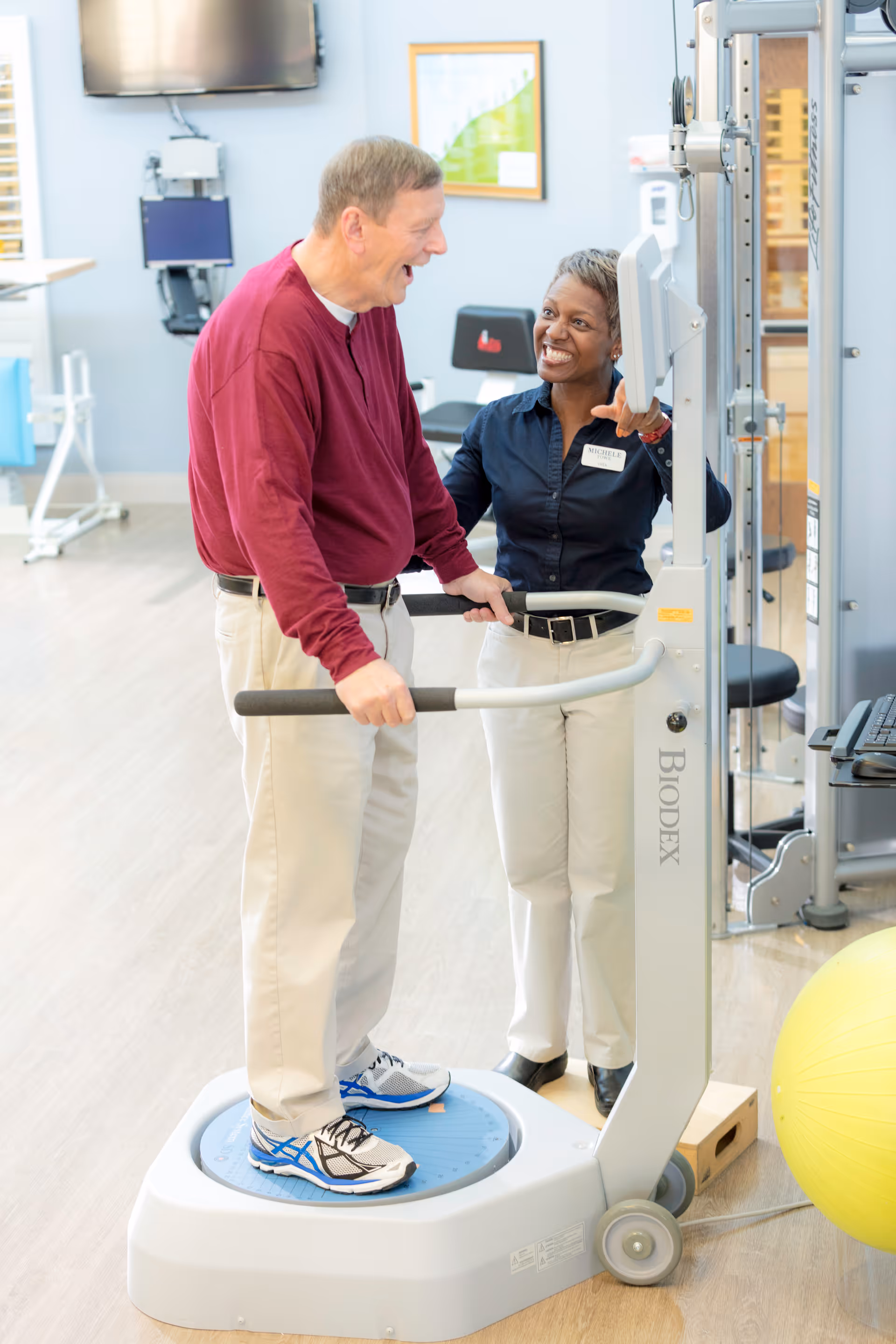 An elderly man stands on a balance exercise machine while a female staff member, wearing a name tag, smiles and assists him in a rehabilitation or physical therapy room with exercise equipment and a large yellow exercise ball visible.