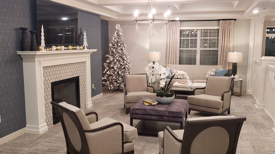 Cozy senior living lounge with armchairs and an ottoman arranged around a fireplace, a decorated Christmas tree, and a sofa under a chandelier.