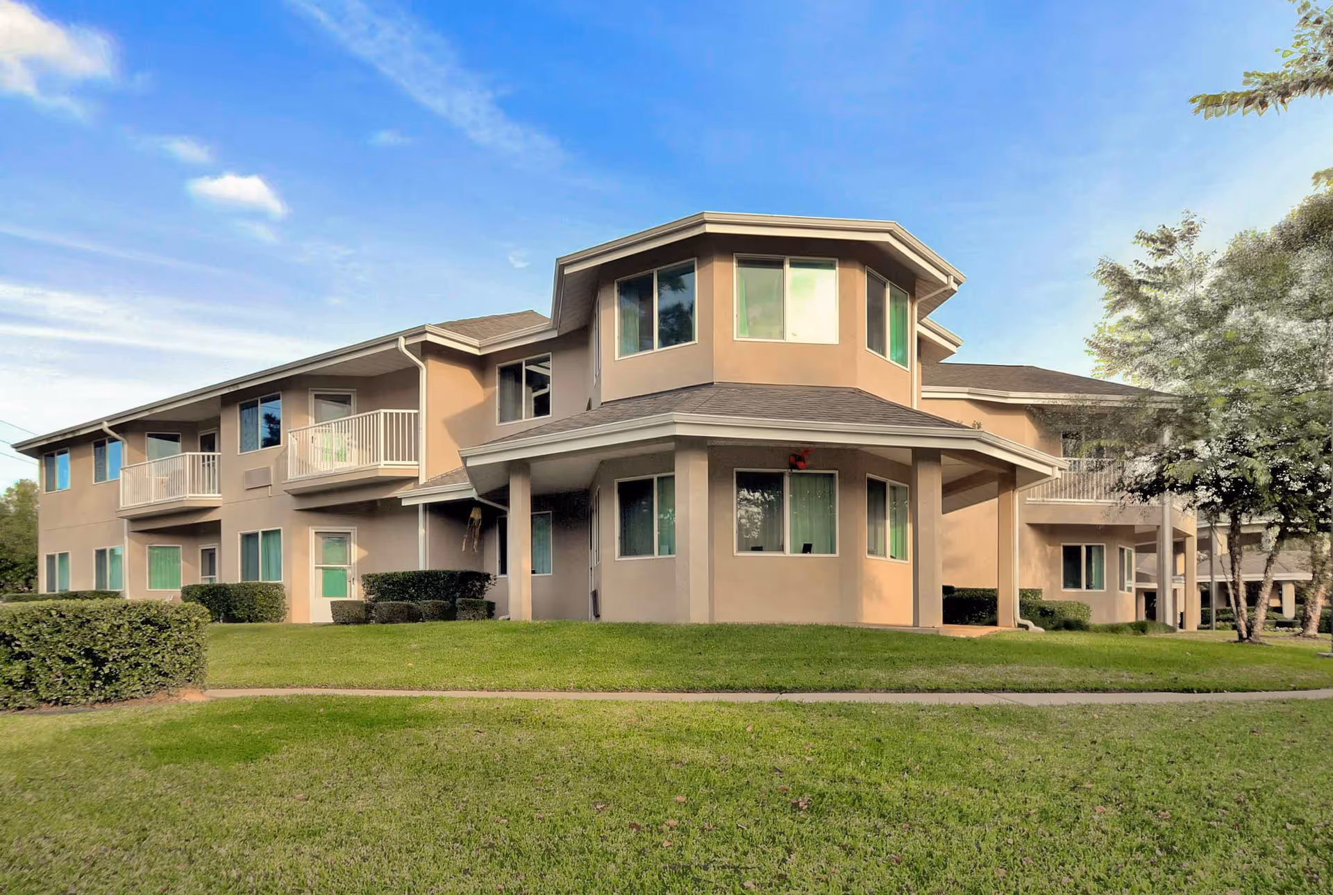 Two-story beige apartment-style building with balconies and many windows set behind a green lawn under a blue sky.