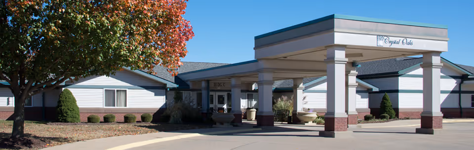 Exterior view of Crystal Oaks senior living facility showing a covered entrance with columns, a driveway, and a tree with autumn-colored leaves on the left side.