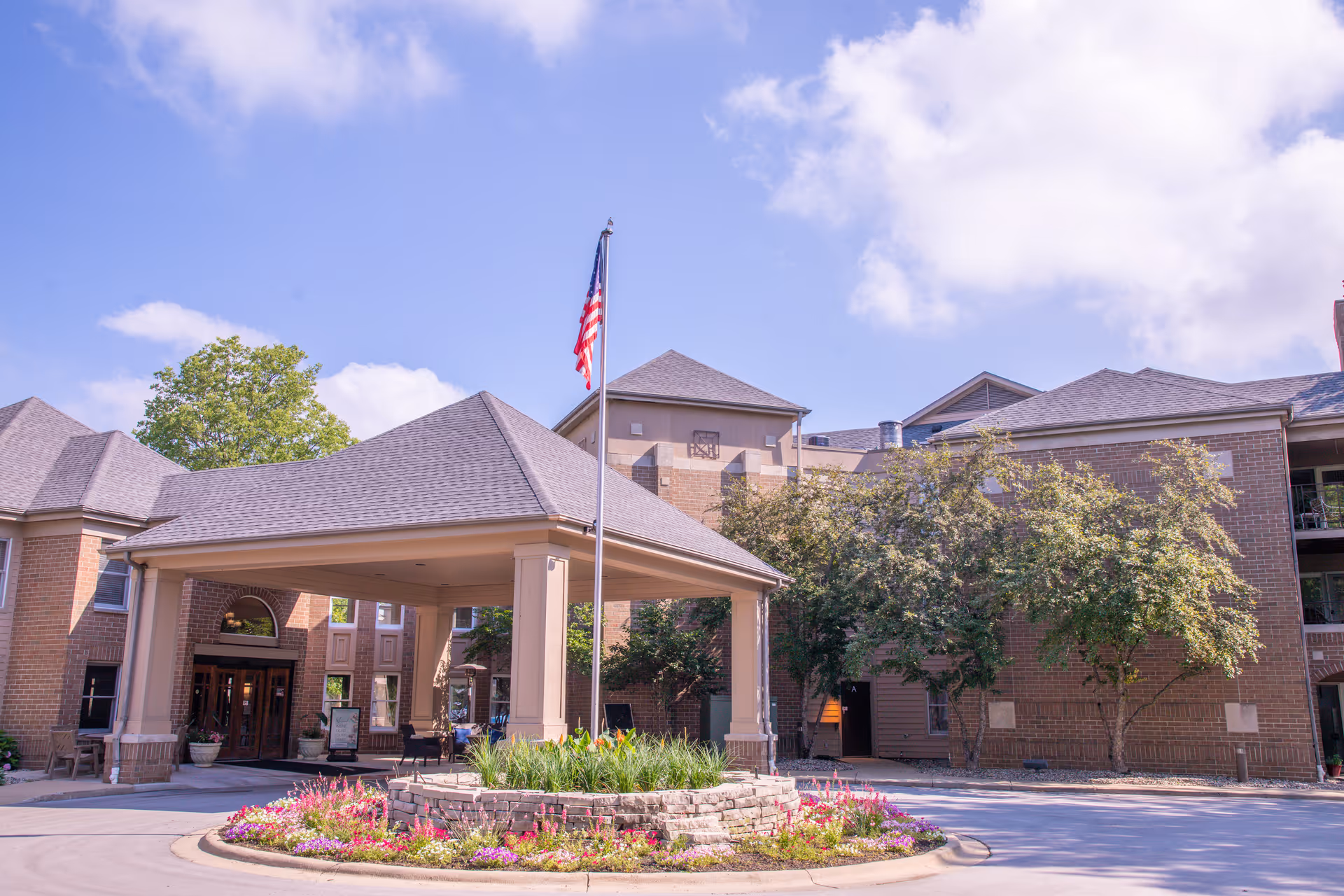 Front entrance of a brick senior living community with a covered porte-cochere, an American flag, and a landscaped roundabout.