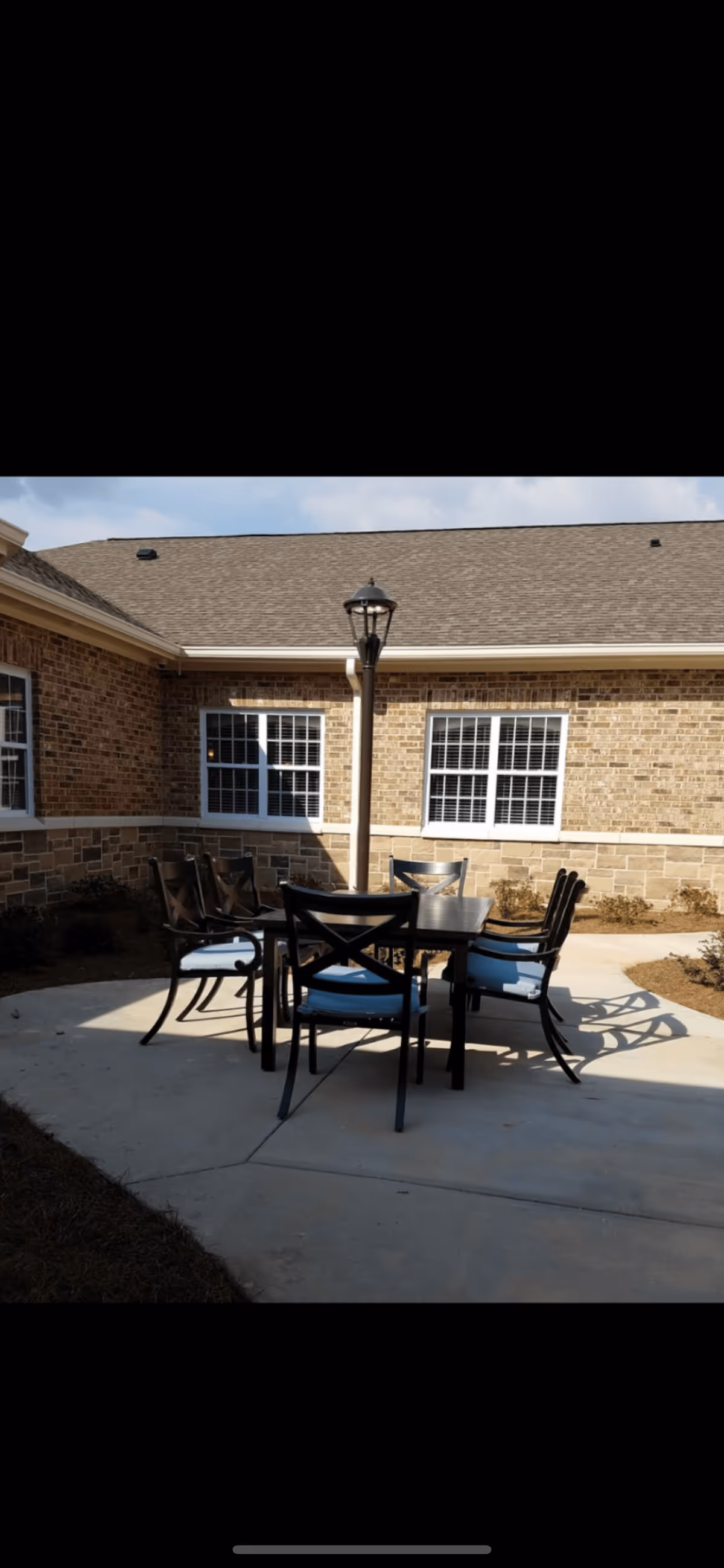 Outdoor patio area with a rectangular table surrounded by six black chairs with blue cushions, situated on a concrete surface. The background shows a brick building with two windows and a lamp post in the center behind the table.
