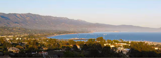 Panoramic coastal view showing a bay with sailboats, shoreline homes, and distant mountains.