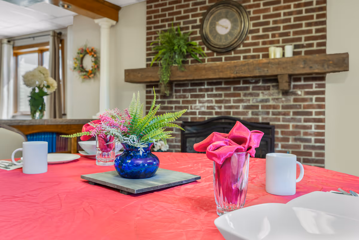 A dining table set with a red tablecloth, white mugs, white plates, and glasses with pink cloth napkins folded inside. A blue vase with green and pink artificial flowers is placed in the center of the table. In the background, there is a brick fireplace with a wooden mantel, a round clock, and some candles. A window with curtains and a floral wreath on the wall are also visible.