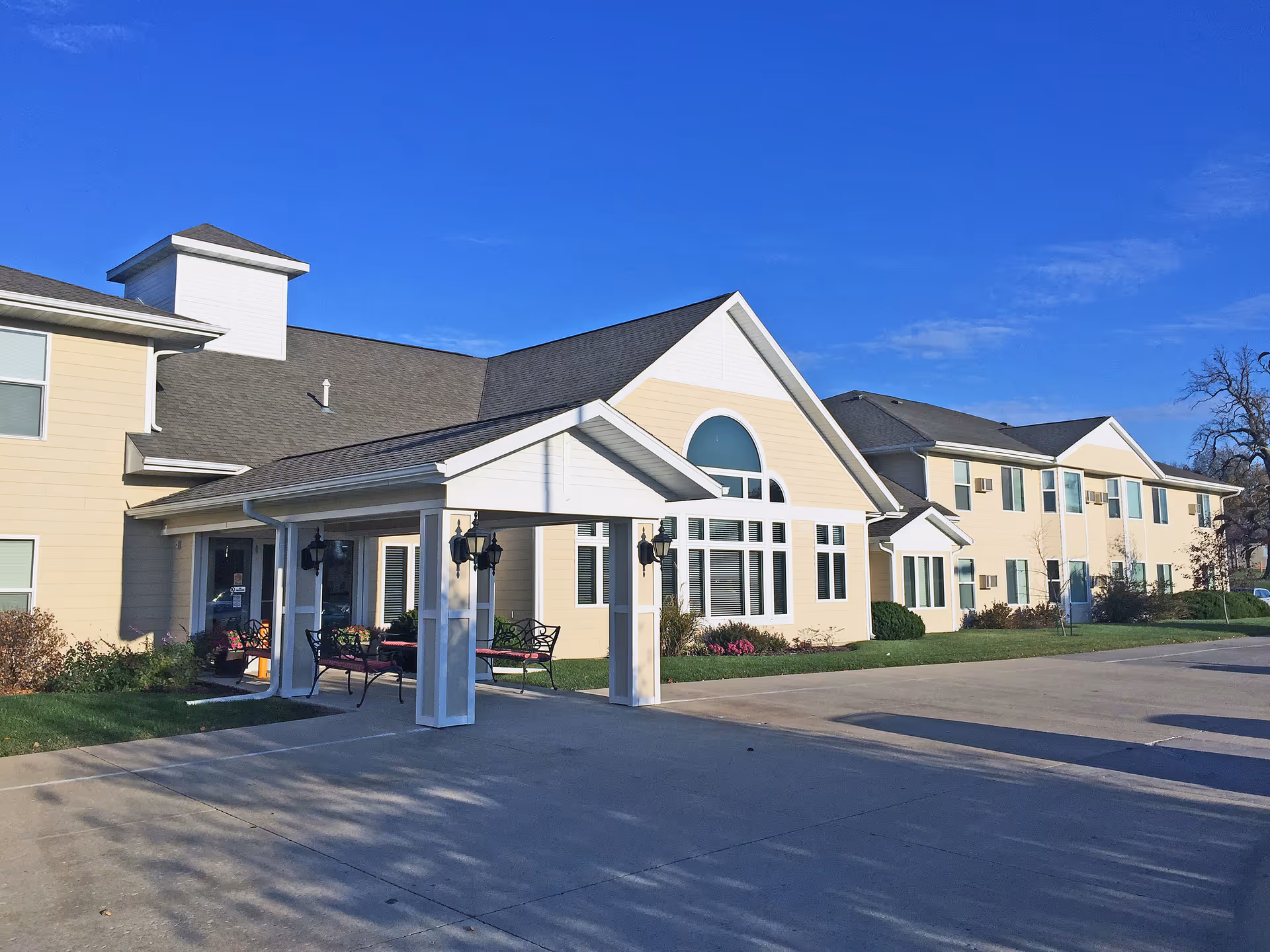 Exterior view of Meadow Ridge Senior Living facility showing a two-story beige building with white trim, large windows, and a covered entrance with benches underneath. The sky is clear and blue.