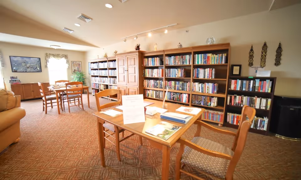A cozy library or reading room with wooden bookshelves filled with books along the walls. There are wooden tables and chairs arranged for reading or studying. The room has carpeted flooring, a window with curtains, and some framed artwork on the walls. The lighting is warm and inviting.