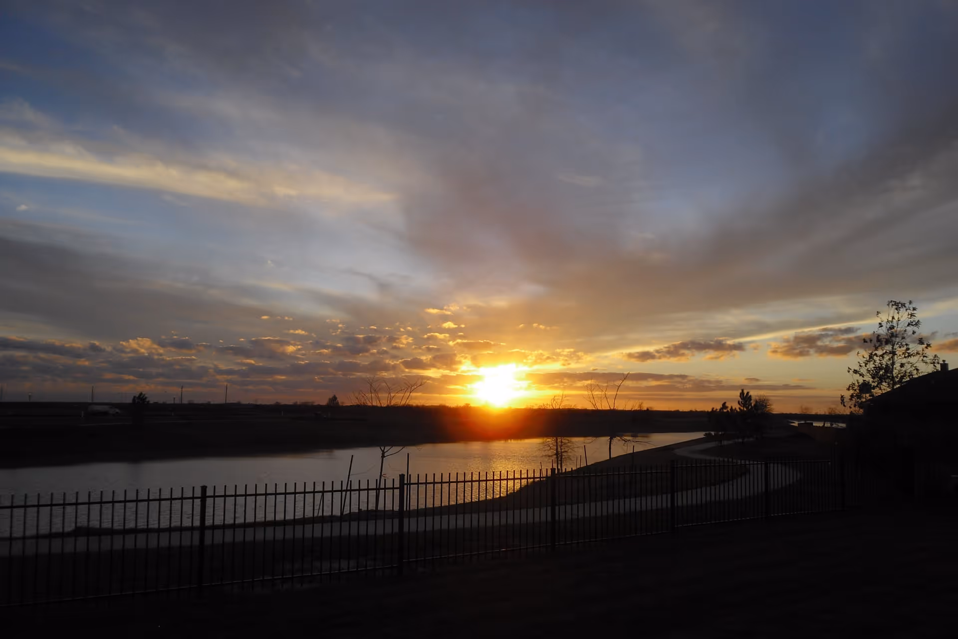 Sunset over a calm lake with a winding paved path along the shore, a black metal fence in the foreground, and scattered trees silhouetted against a colorful sky with clouds.
