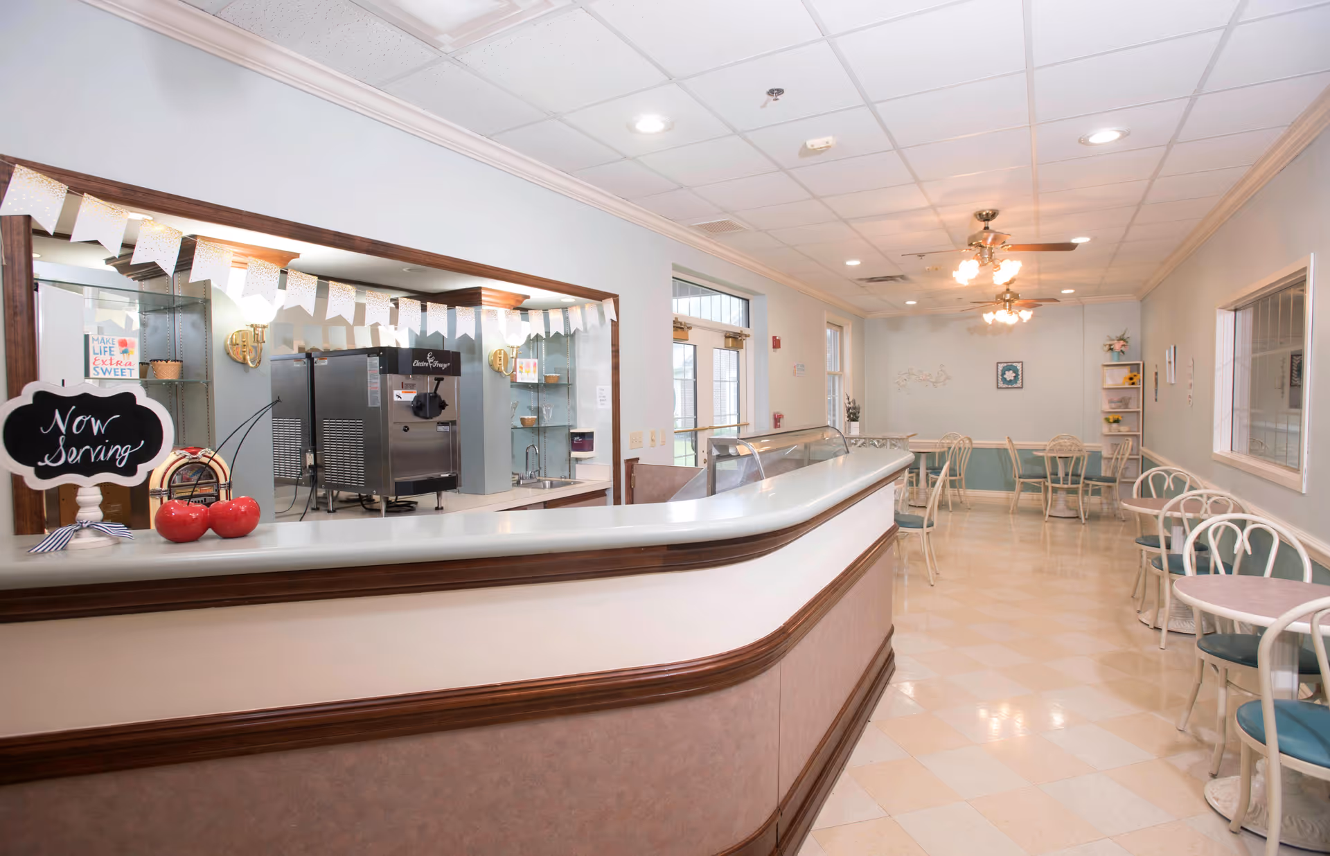 Interior view of a dining area with a long serving counter on the left side. The counter has a small chalkboard sign that reads 'Now Serving' and two decorative red apples. Behind the counter, there is a soft serve ice cream machine and shelves with small decorations. The dining area has several small round tables with chairs arranged along the right side and at the far end of the room. The ceiling has recessed lighting and ceiling fans with lights.