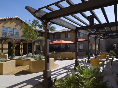 Outdoor courtyard area of a senior living facility with a wooden pergola casting shadows over tables and chairs. There are red umbrellas providing shade, desert landscaping with plants and small trees, and a two-story building with windows surrounding the courtyard.