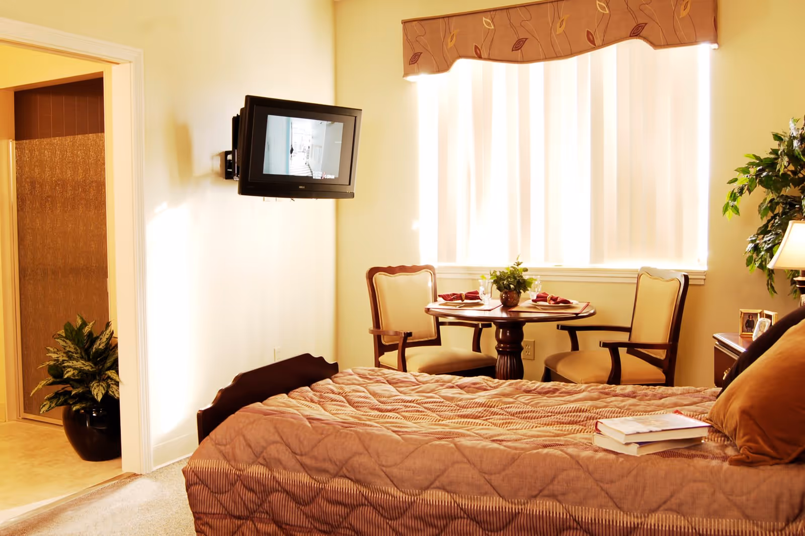 Sunlit bedroom with a bed, wall-mounted TV, a small round table with two chairs, and a doorway to a bathroom.