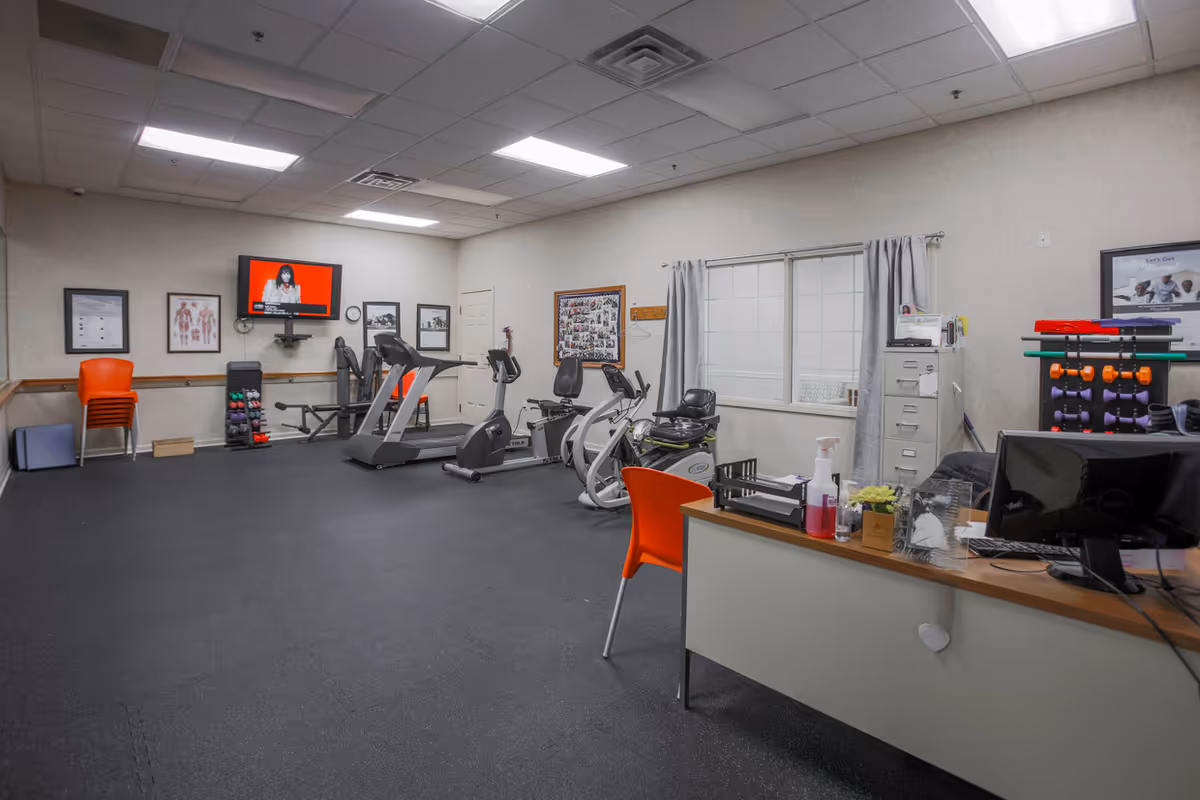 Interior view of a fitness room with exercise equipment including treadmills, stationary bikes, and weights. There is a desk with an orange chair in the foreground, a TV mounted on the wall showing a person on a red background, and various framed pictures and posters on the walls. The room has a window with gray curtains and a filing cabinet next to it.