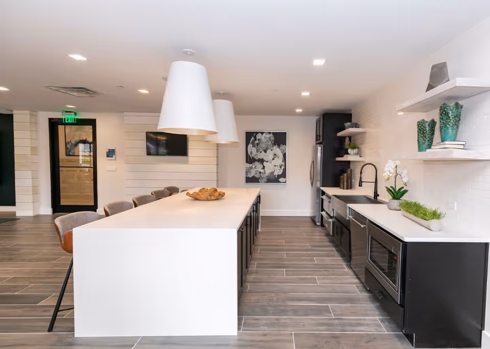 Modern communal kitchen with a long white island, bar stools, pendant lights, stainless appliances and open shelving.