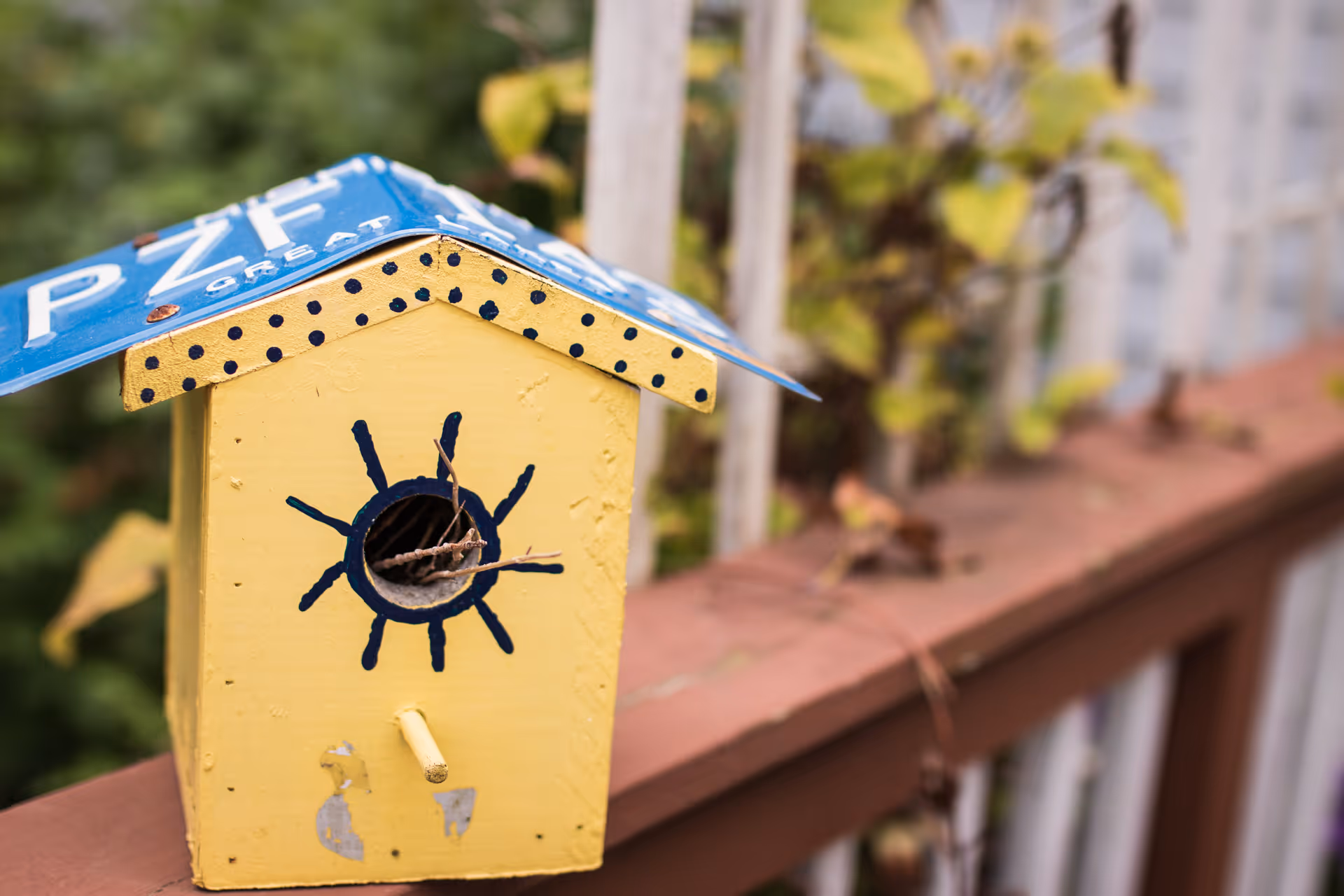 A small yellow birdhouse with a blue roof made from a repurposed license plate, decorated with black dots and a sun design around the entrance hole, mounted on a wooden railing with blurred green foliage in the background.