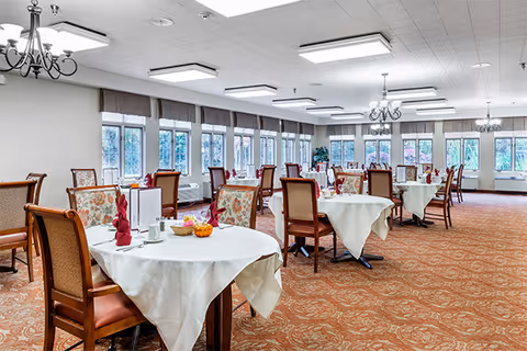 A spacious dining room with multiple round tables covered with white tablecloths. Each table is set with folded red napkins, small decorative pumpkins, and dining utensils. The room has large windows along one wall letting in natural light, patterned carpet flooring, and chandeliers hanging from the ceiling.
