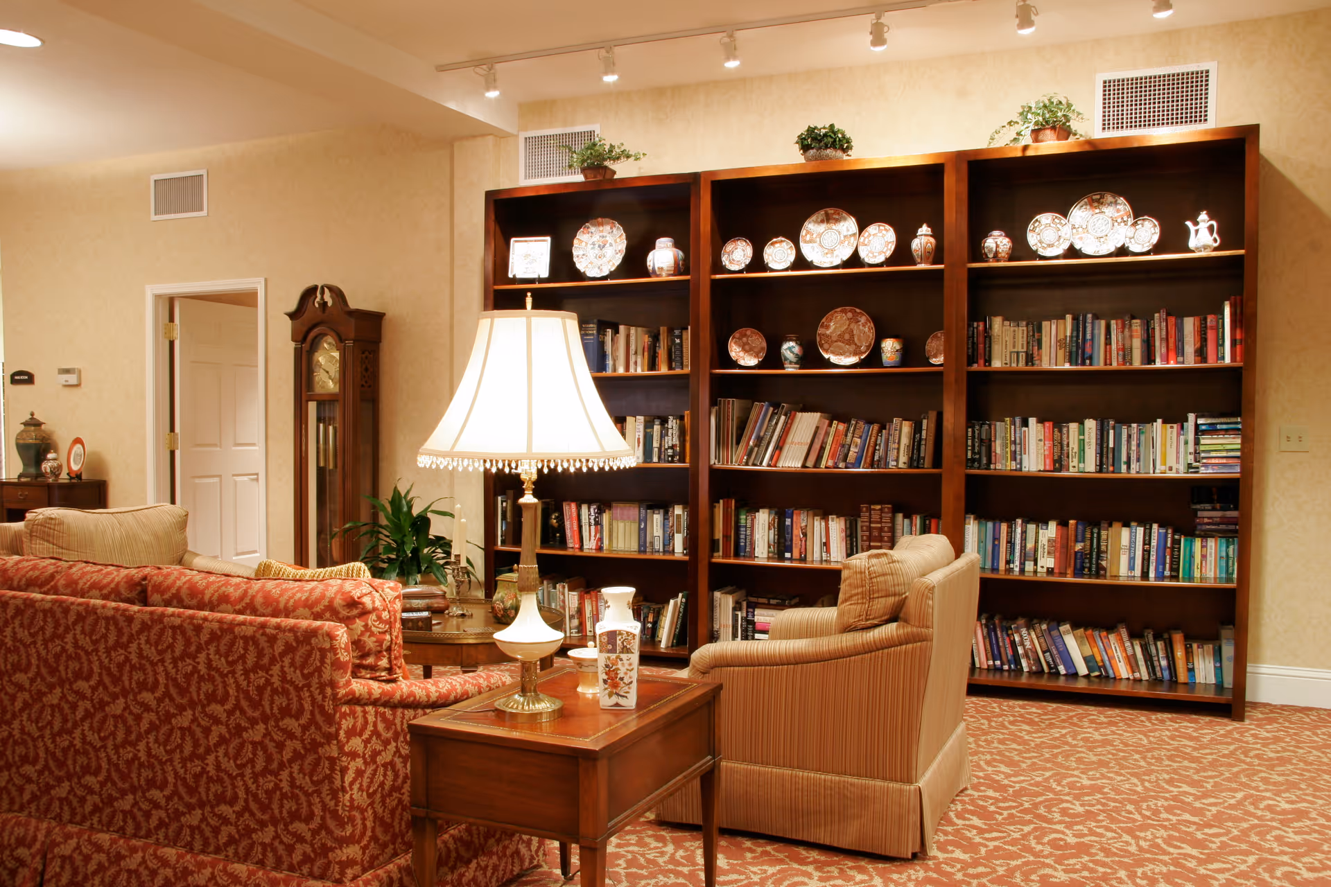 Cozy seating area with upholstered chairs and sofa facing large wooden bookshelves filled with books and decorative plates, lit by table lamps.